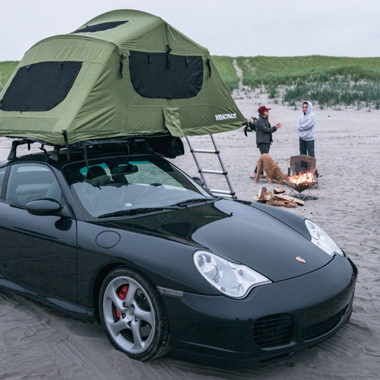 A black 2004 Porsche 911 with a green Yakima rooftop tent owned by Brock Keen of Instagram handle @996roadtrip sitting on a beach with two women in the background next to a campfire