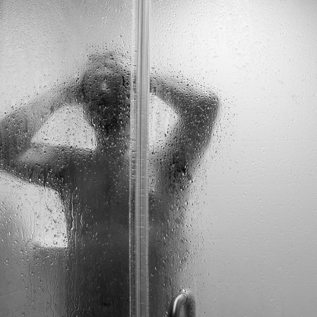 A black and white photo of a person showering, as soon through a shower door