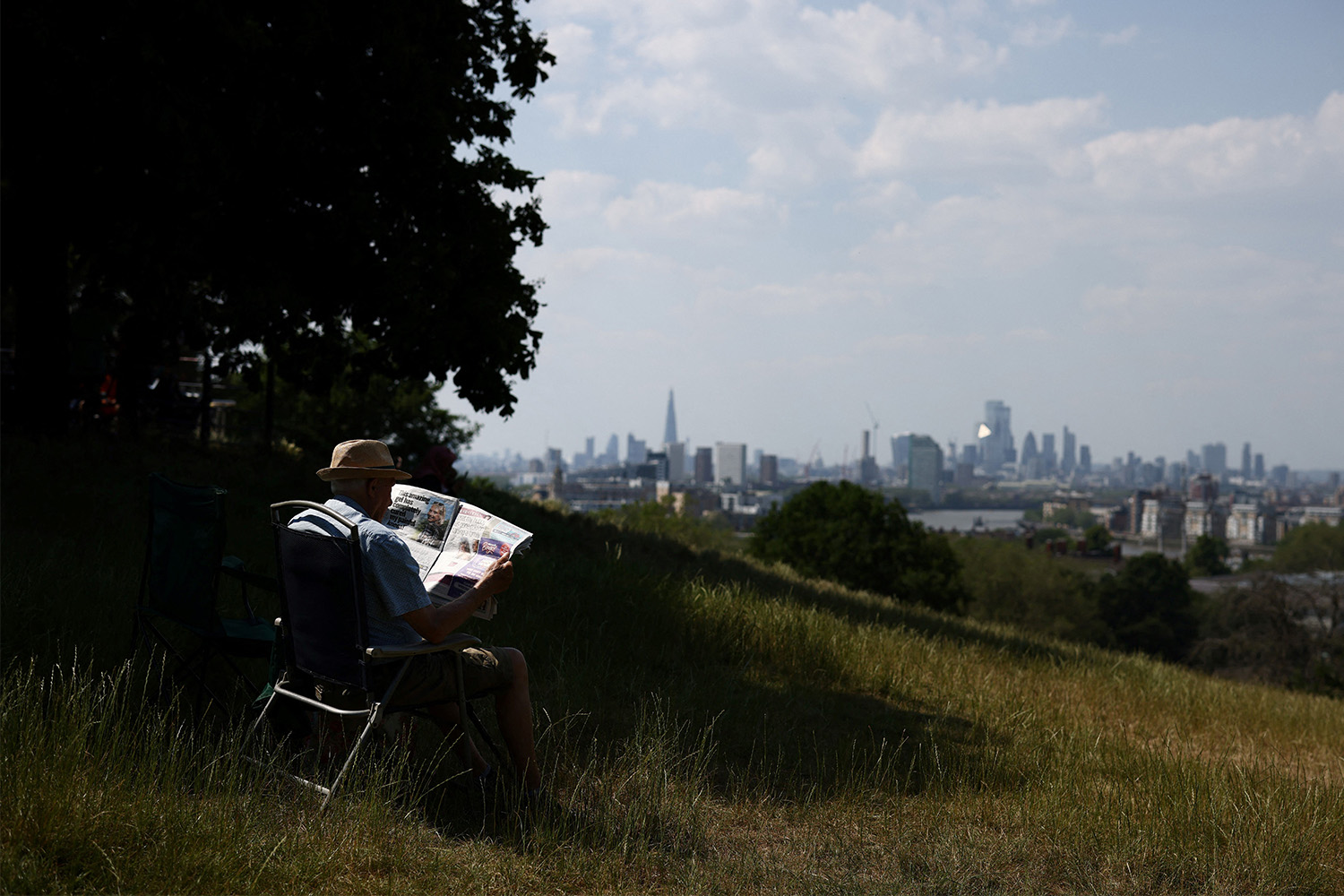 A man reading a newspaper on a grassy hill, with a city in the distance.