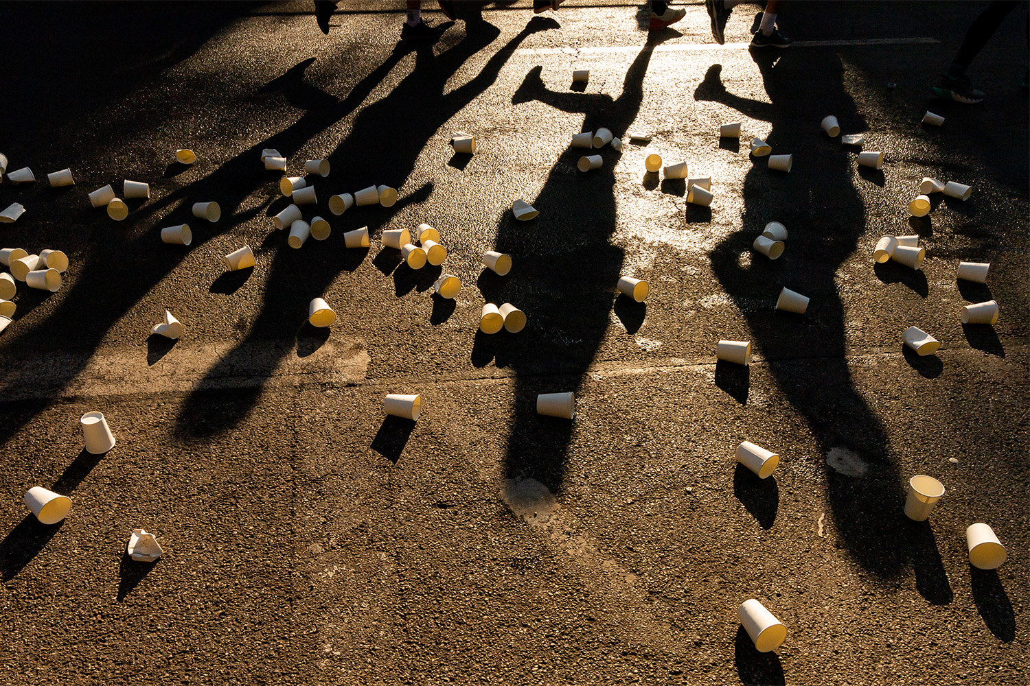 A bunch of marathon cups on the concrete, with runners' shadows in the background.