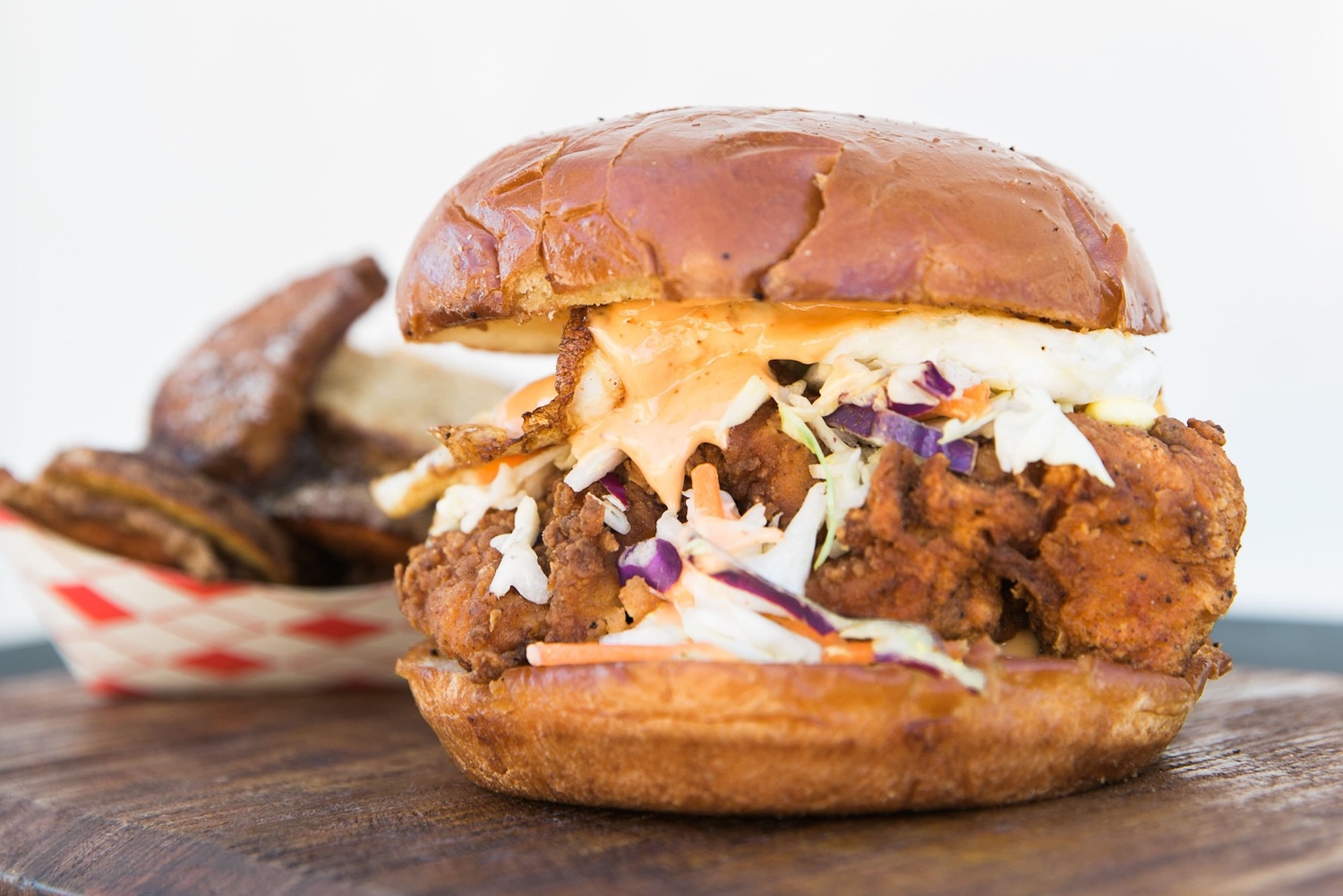 a fried chicken sandwich topped with slaw on a wooden cutting board with fries in the background