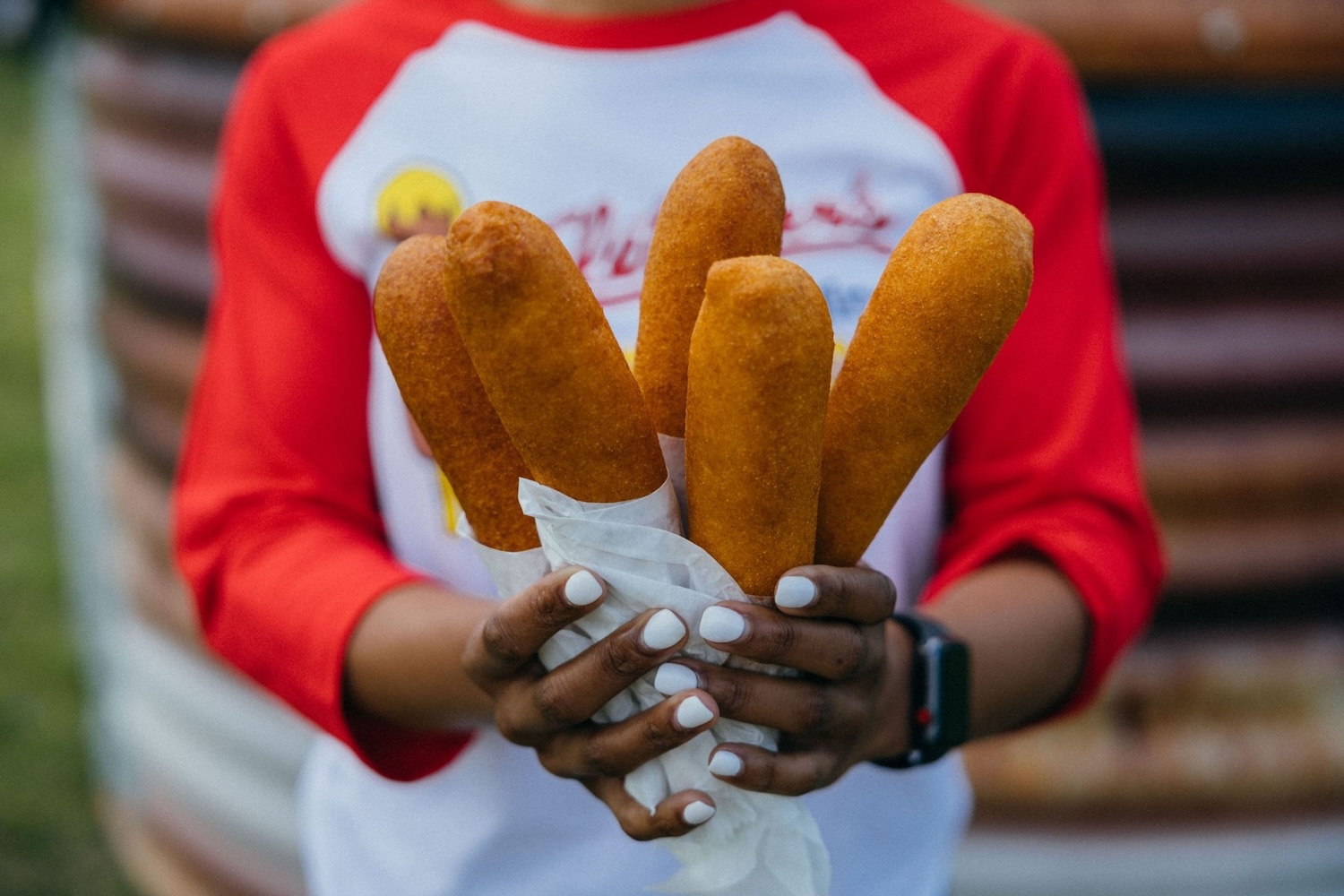 person holding 5 corndogs in their hand, nails painted white, red and white shirt on