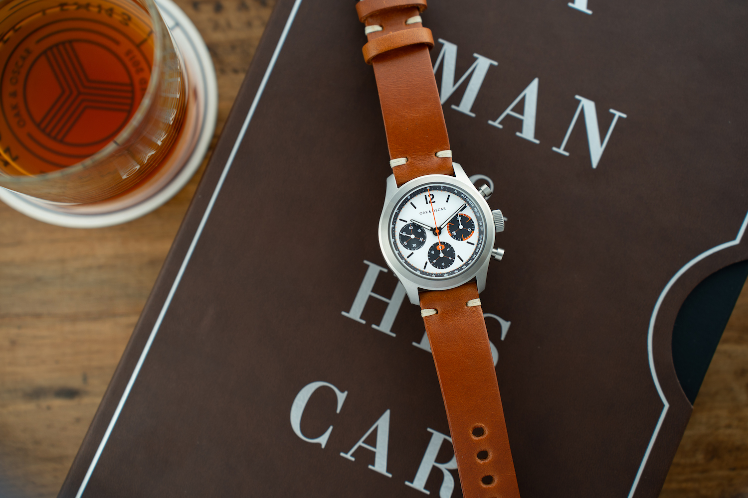 Watch with brown leather strap on top of a book next to a whiskey glass on a table.