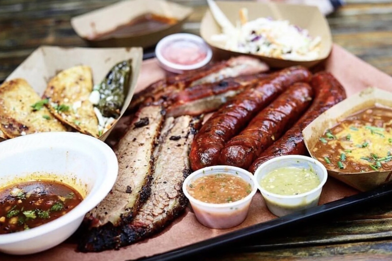 a tray of bbq food with brisket, sausage, sauces, beans and birria tacos