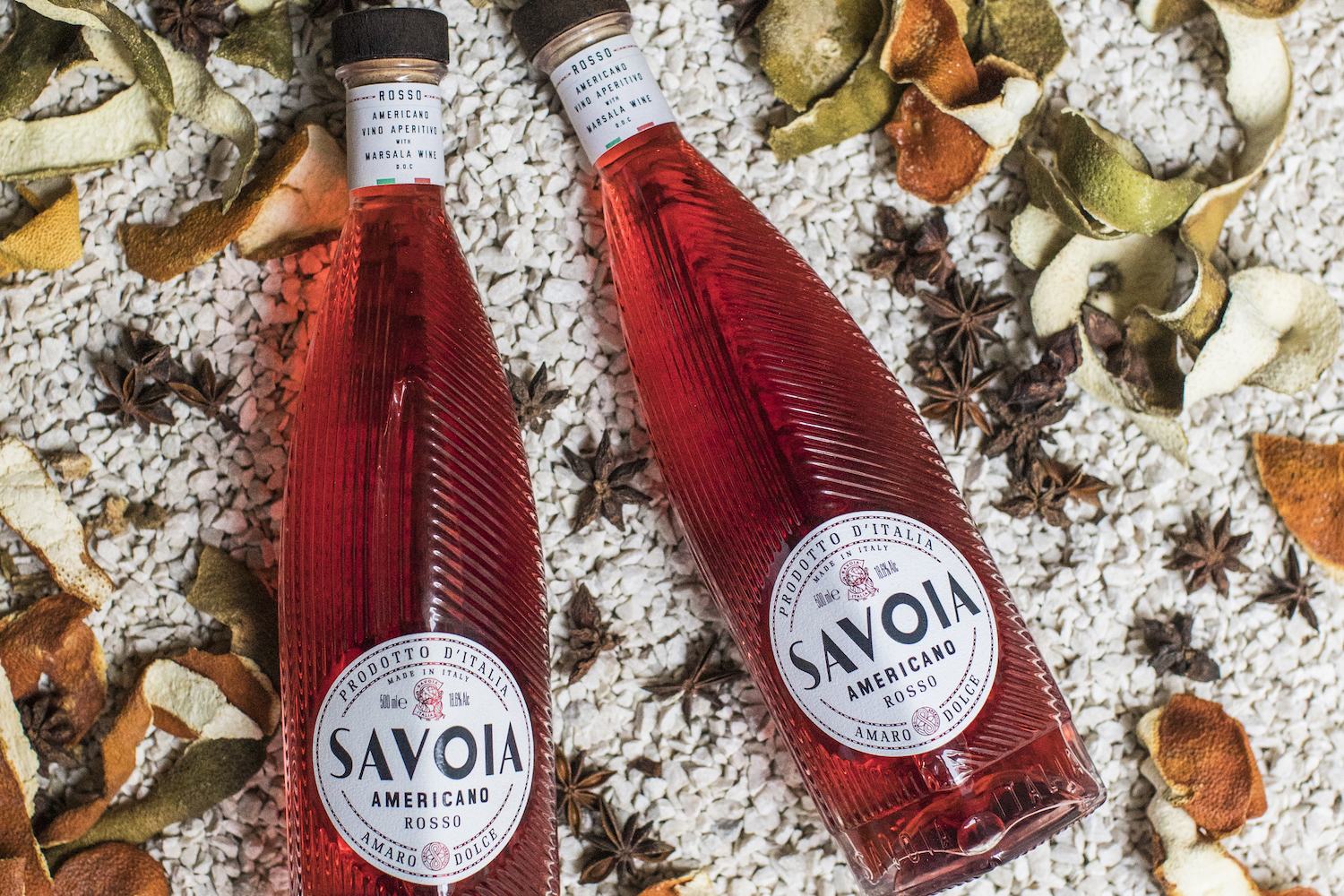 two bottles of Savoia Americano Rosso laying on white stones surrounded by citrus peels and star anise