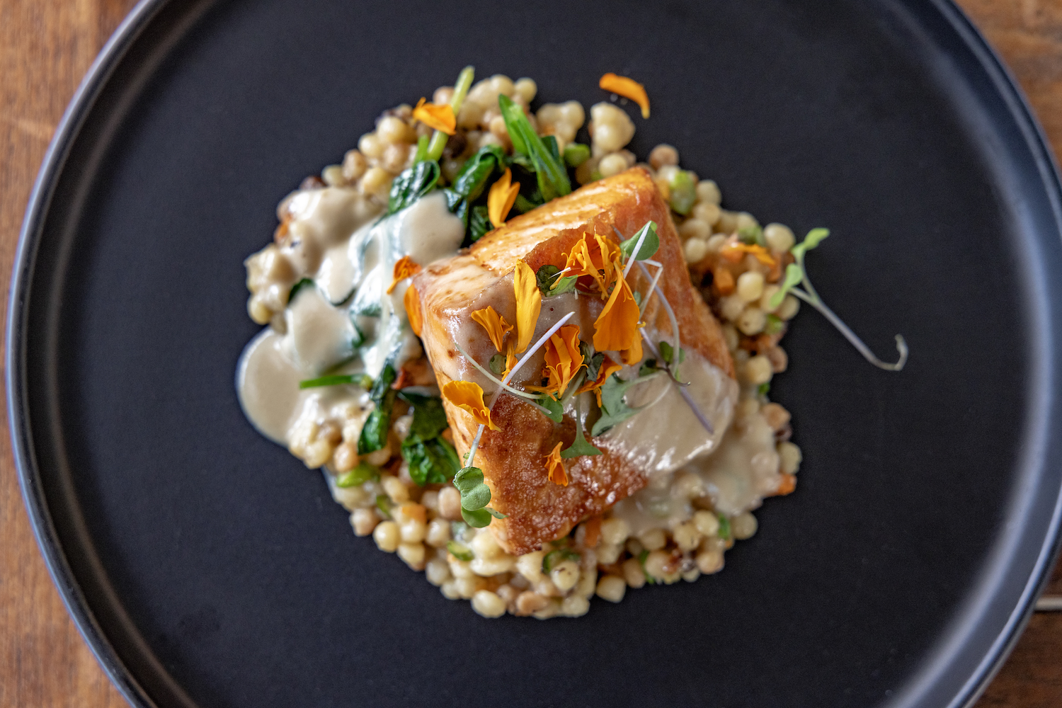 a black plate of roasted salmon and fregola with edible flowers