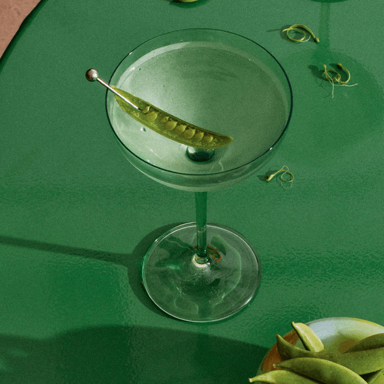 a martini garnished with a snap pea sitting on a green table