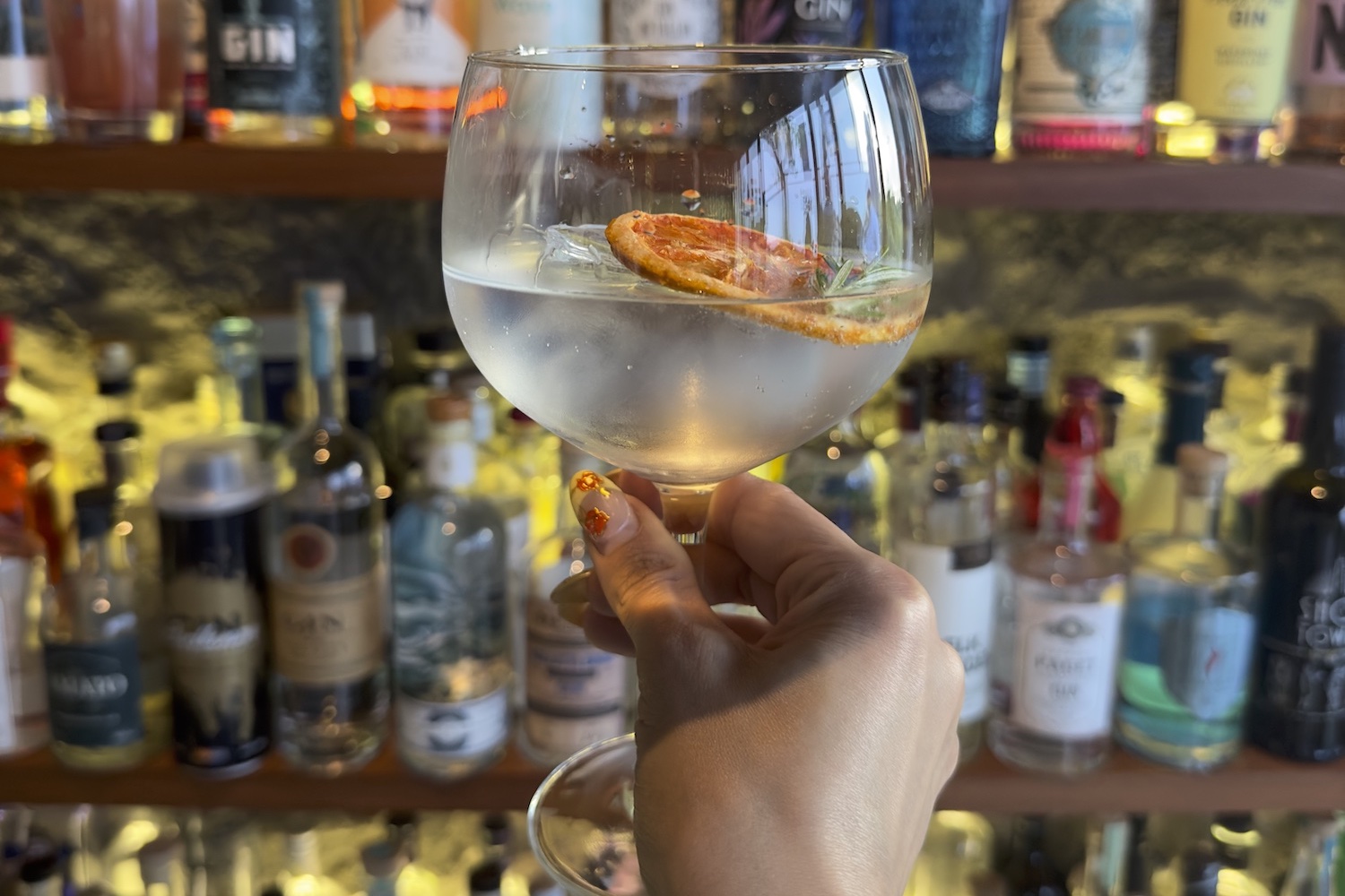 a hand holding a gin and tonic in front of a wall of gin bottles