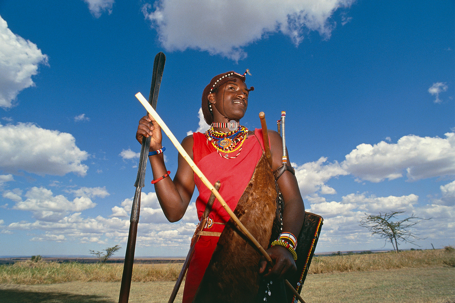 A Maasai warrior holding his spear, the dry savanna spread out behind him.