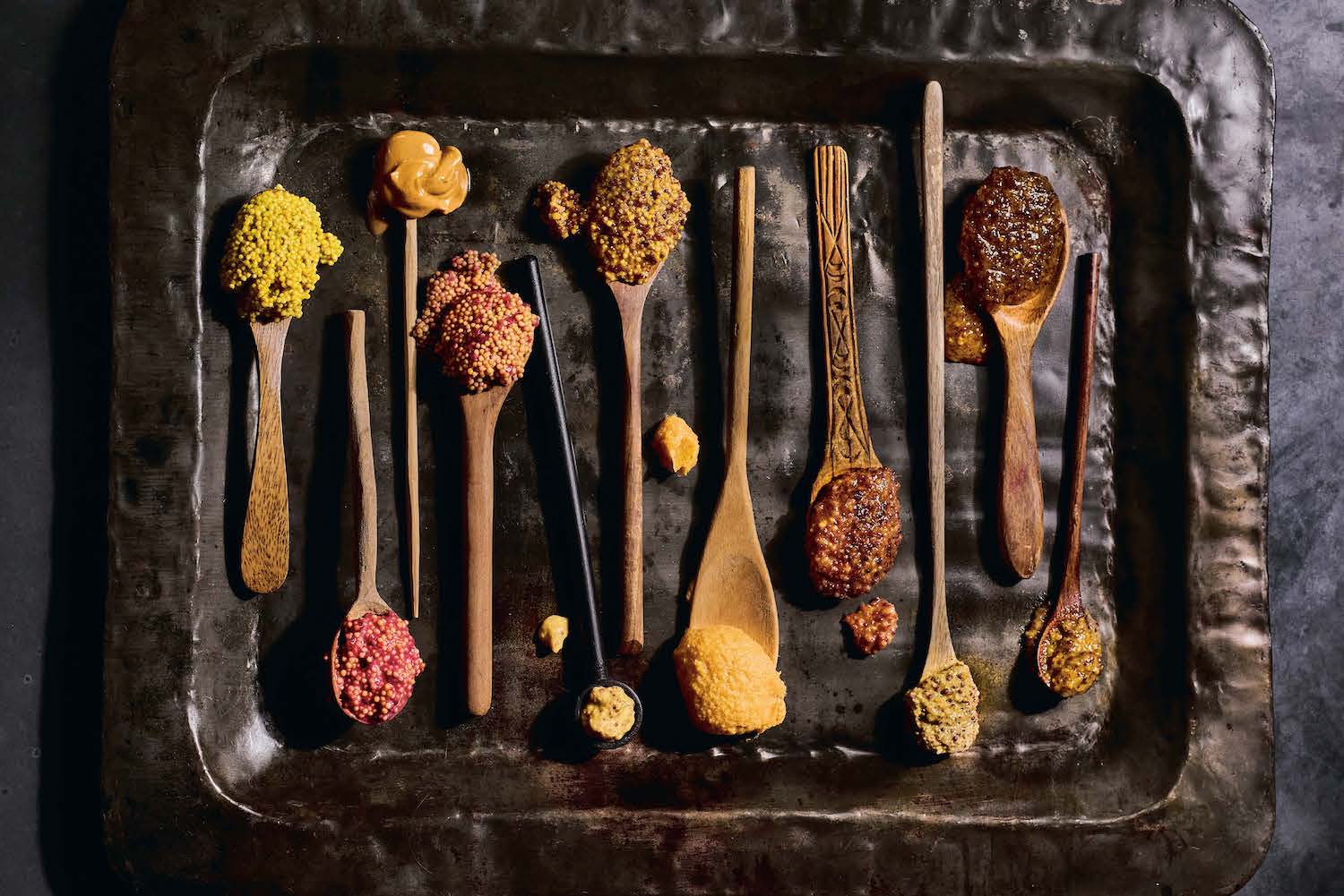 wooden spoons holding different types of mustard on a tin tray