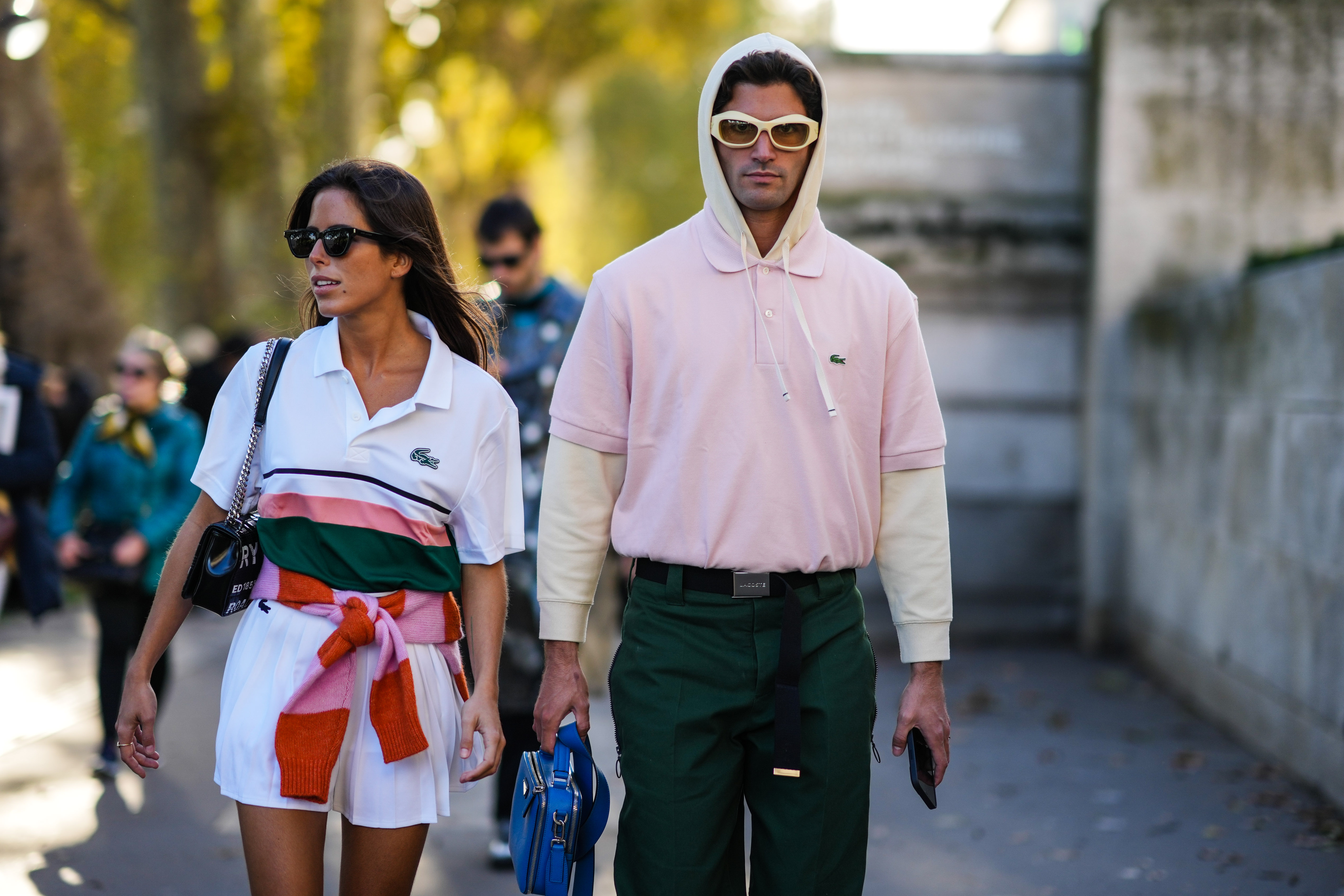 PARIS, FRANCE - OCTOBER 05: A guest (L) wears sunglasses, a white with dark green and pink straps polo from Lacoste, a pale pink and orange striped wool pullover, a white pleated / accordion short skirt, a black shiny leather shoulder bag, a guest (R) wears sunglasses, a pale pink polo shirt from Lacoste, a pale yellow hoodie sweater, black belt from Lacoste, dark green sport pants, a blue shiny leather handbag, outside Lacoste, during Paris Fashion Week - Womenswear Spring Summer 2022, on October 05, 2021 in Paris, France.