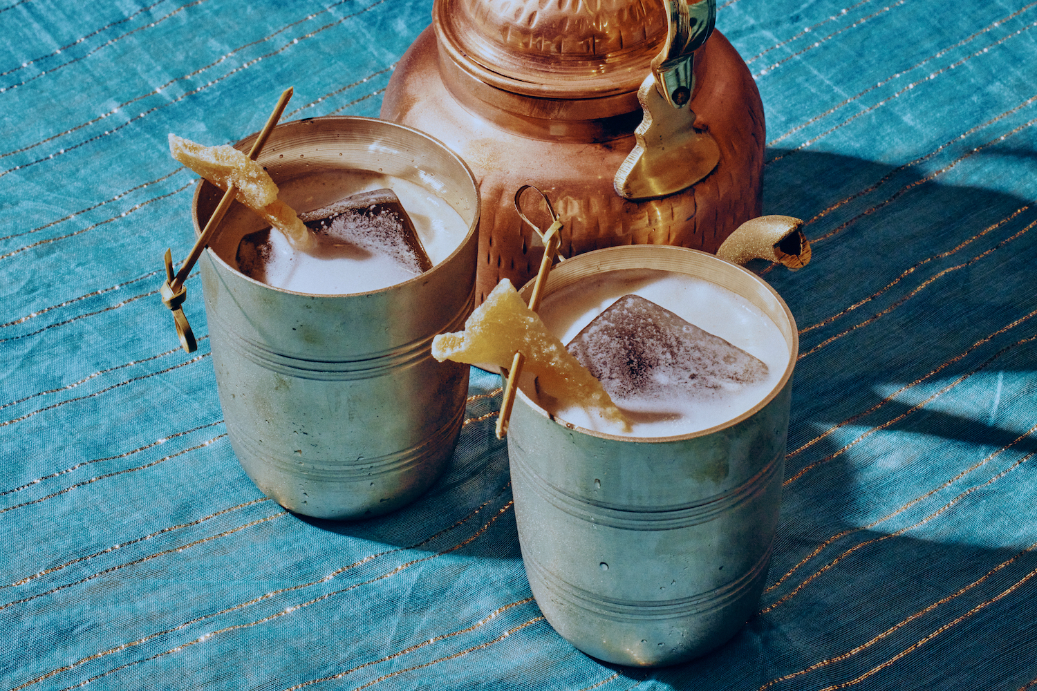 two cocktails in mule mugs garnished with candied ginger next to a teapot on a blue and gold tablecloth