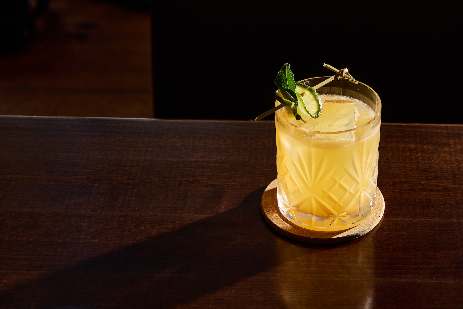 a yellow cocktail in a rocks glass on a wodden table, garnished with lime and mint