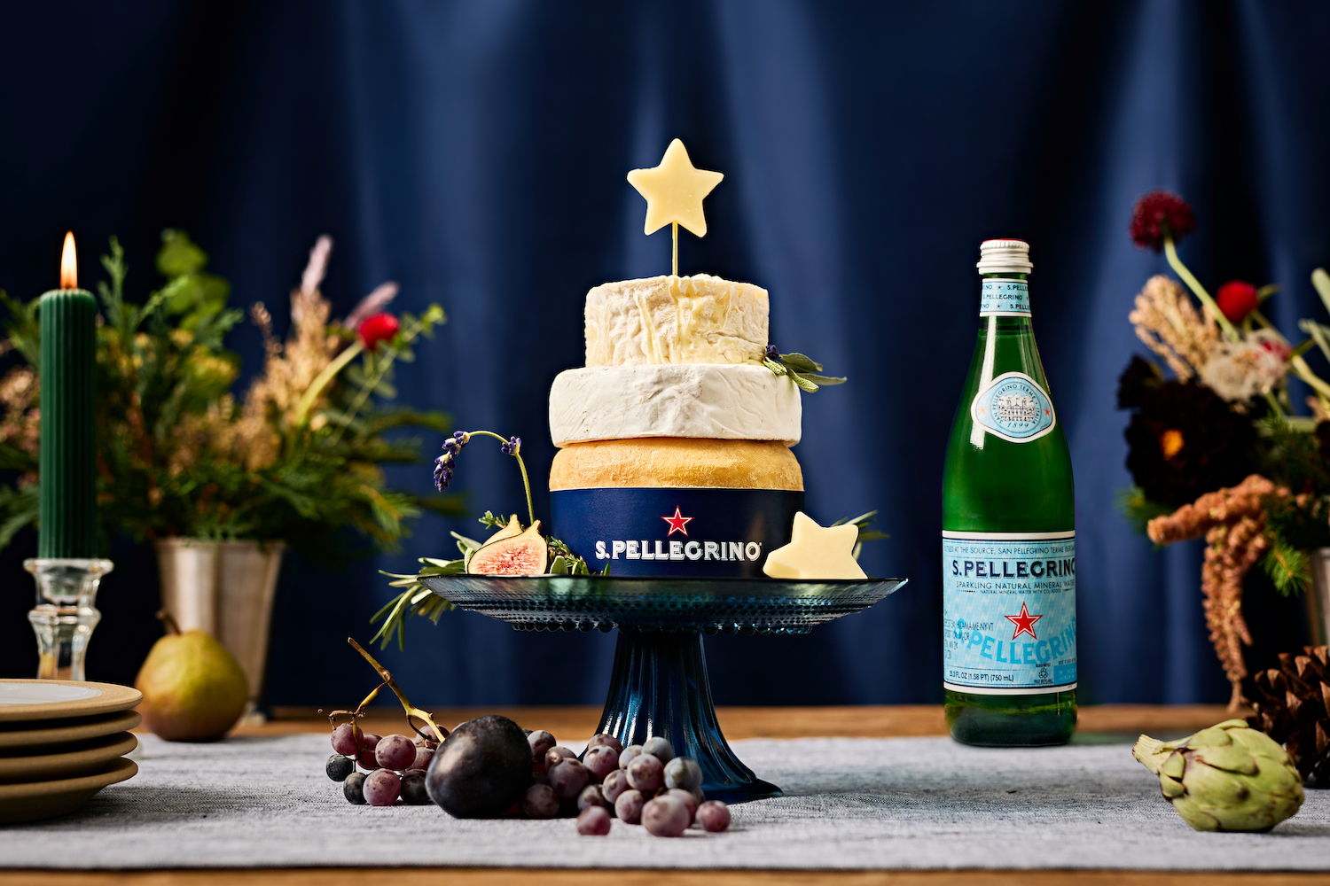 a tier of three round cheeses topped with a cheese star on a blue cake stand sitting on a table with a candle, flowers and S.Pellegrino