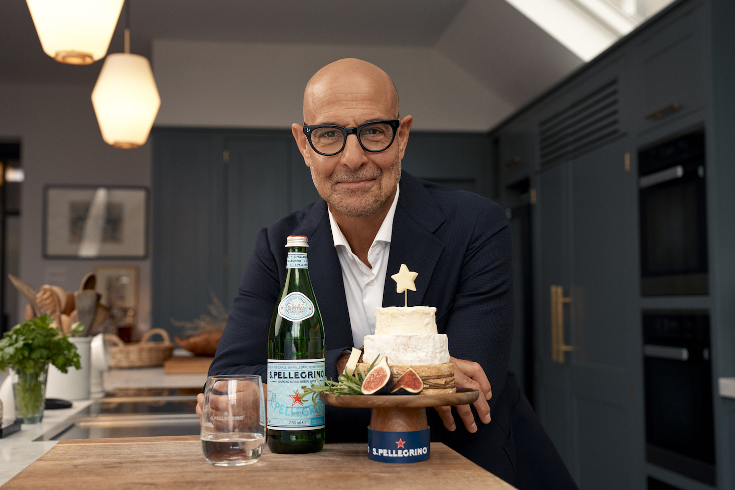 stanley tucci wearing a navy suit standing behind a wood-topped kitchen island with a bottle of S.Pellegrino and a tower of cheese