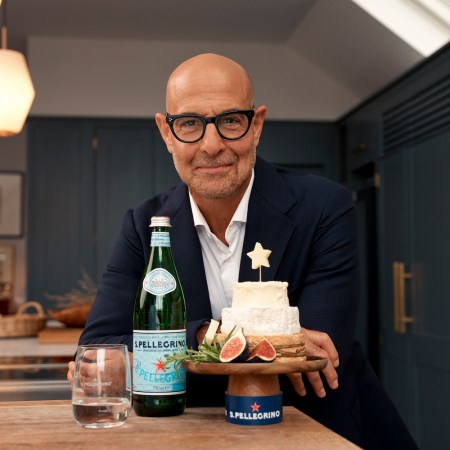 stanley tucci wearing a navy suit standing behind a wood-topped kitchen island with a bottle of S.Pellegrino and a tower of cheese