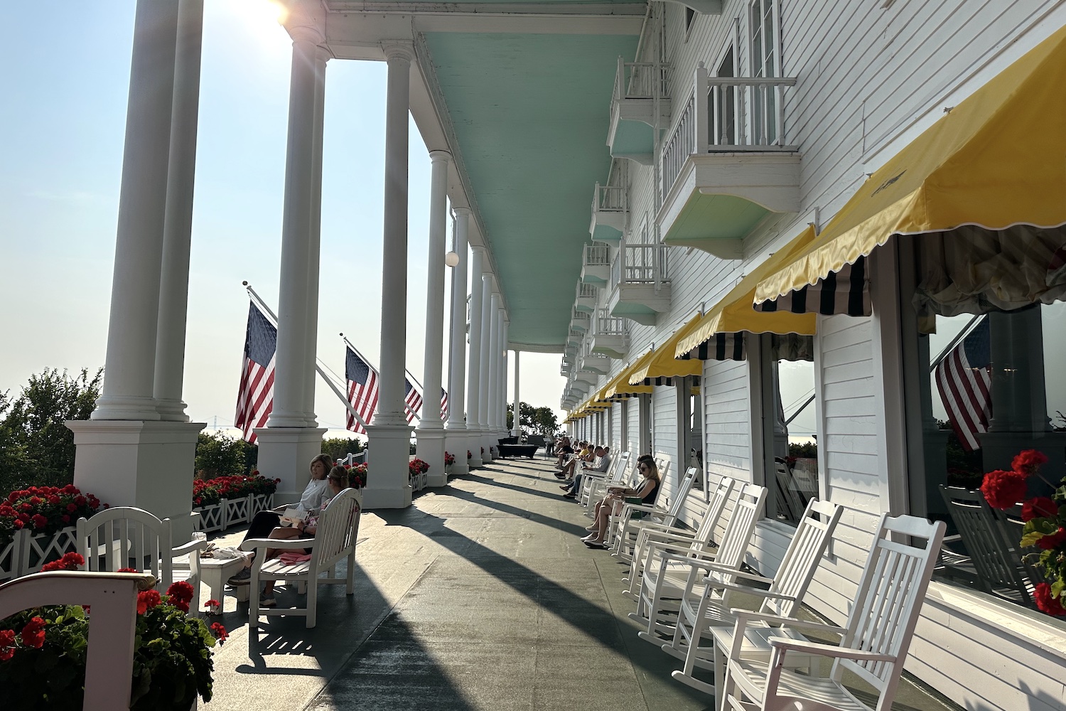 a long porch with american flags, white columns, yellow awnings and white rocking chairs