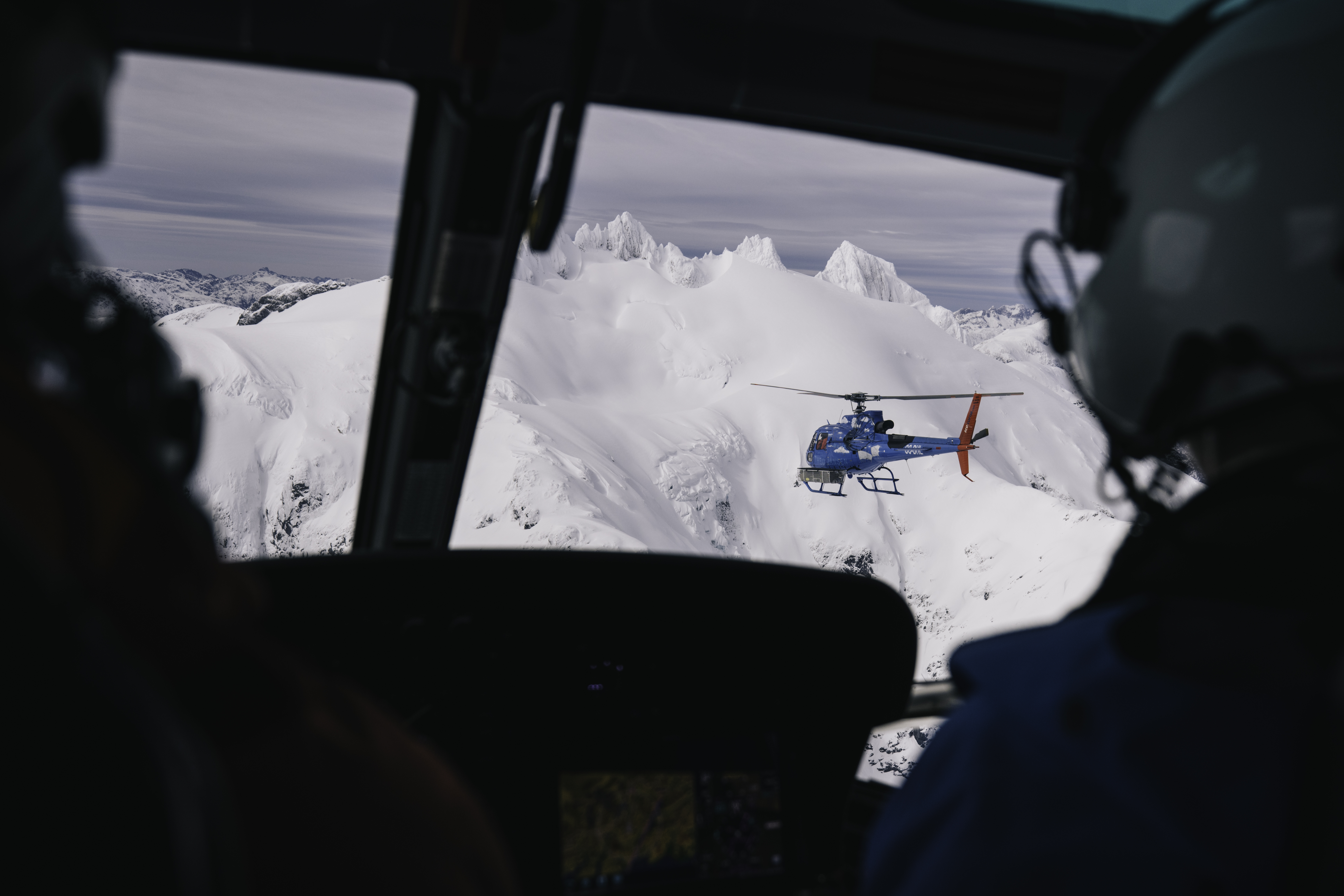 A view of another helicopter from the cockpit of a helicopter, skimming over snowy mountains in the Andes.