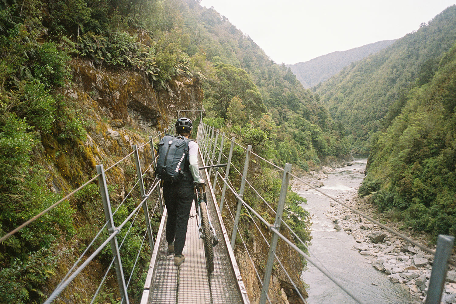 A female cyclist pushes her bike across a pedestrian suspension bridge.