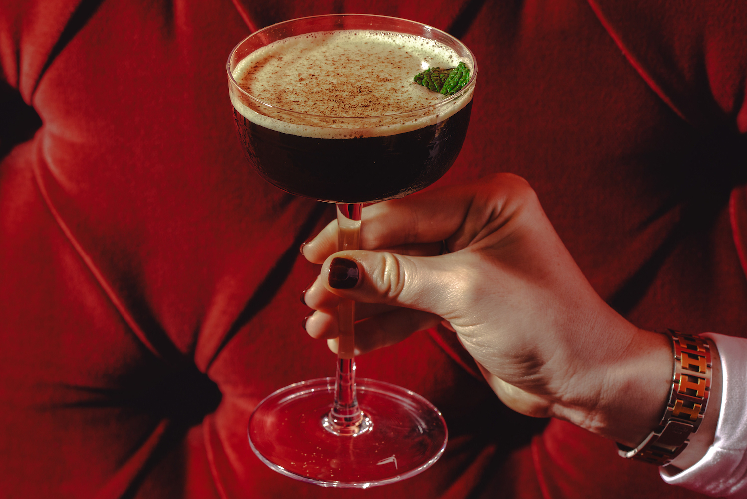 a woman with dark red nails holding an espresso martini in front of a red couch