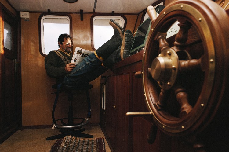 A man relaxes in the cabin of a boat, reading a magazine with his feet up on the control panel, embracing an offline moment.