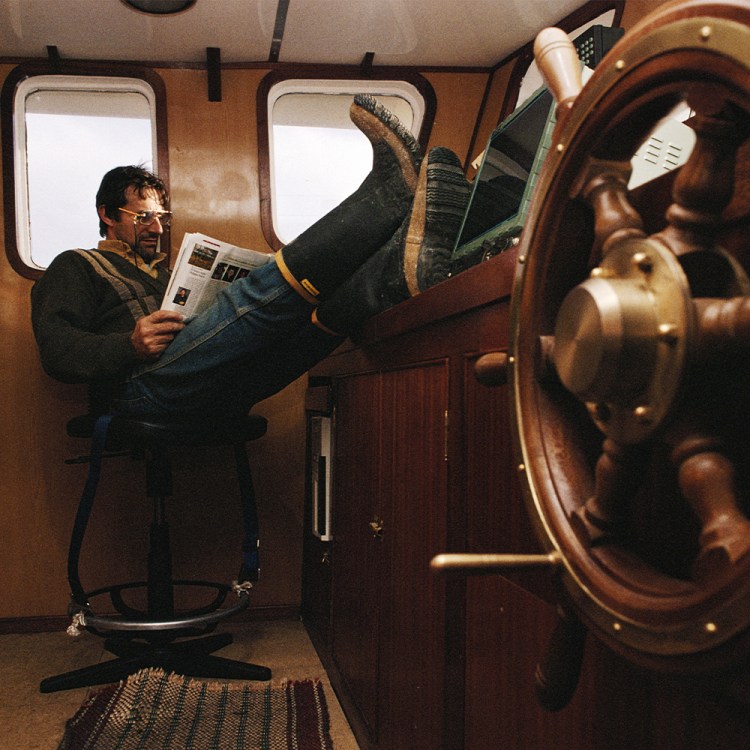 A man relaxes in the cabin of a boat, reading a magazine with his feet up on the control panel, embracing an offline moment.