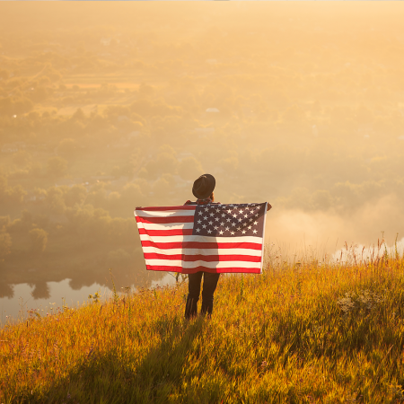 a man standing on a hill at golden hour holding an american flag behind his back