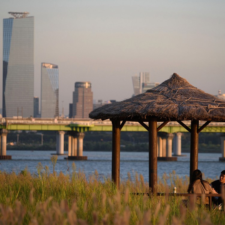 A young couple enjoys a quiet moment under a thatched-roof gazebo by the Han River in Seoul, with the modern skyline in the background, embodying the philosophy of sohwakhaeng—finding happiness in small, simple moments.